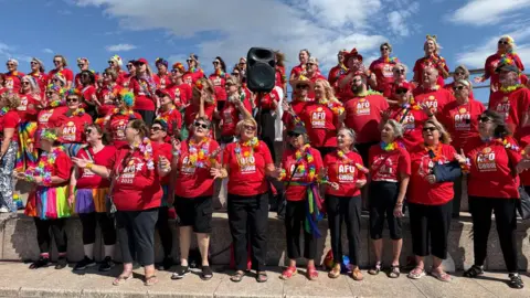 About 50 people in red tops and rainbow leis round their necks. They are mid- song.