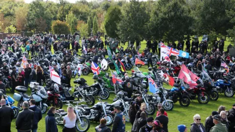 National Memorial Arboretum People gathered at Ride to the Wall. There are many bikes and a large crowd of people. There are also different types of flags. 