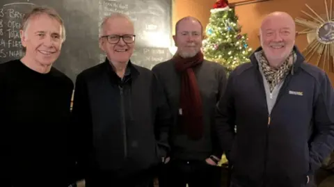 Four men from the band DNA are standing smiling at the camera - there is a Christmas tree and blackboard in the background. 