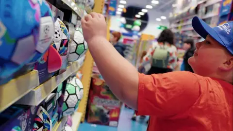 Make-A-Wish UK Jaxen is pictured in a bright orange T-shirt and blue peaked cap. He is reaching up to choose a football from a shelf in a toy shop. 