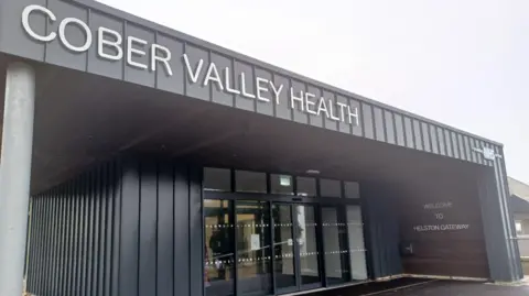 The front of a grey building with automatic doors. It says ' Welcome to Helston Gateway' on it and 'Cober Valley Health'. The sky is grey. 