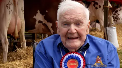 A grey haired man in a blue shirt with a red, white and blue rosette on his chest sits in front of dairy cows