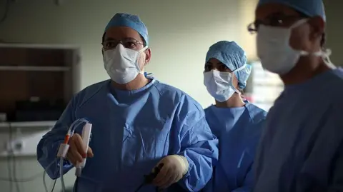 A surgeon and his theatre team perform key hole surgery to remove a gallbladder at the Queen Elizabeth hospital in Birmingham. The doctors are all wearing blue overalls and white masks and staring off camera. The main surgeon is holding a medical device.