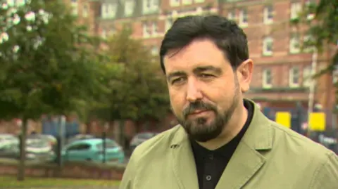 Paul Doherty stands in front of the Royal in a black shirt and a green coat, with black hair and beard