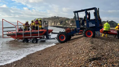 A rescue boat with a crew of four in hi vis and helmets on a pebbly shore attached to a towing vehichel 