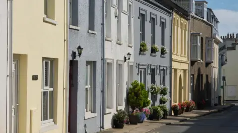 A row of houses on High Street in Port St Mary with the carriageway running alongside the pavement outside them. they are painted yellow, white and grey and some have flower pots and window baskets outside.