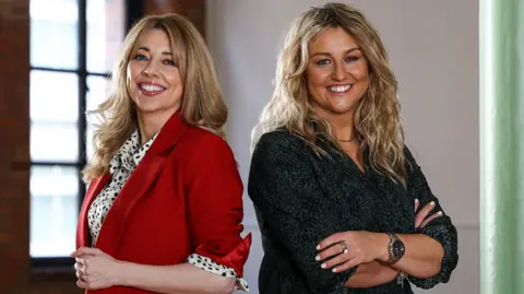 Anna Curran and Eve Blair are standing back to back with their arms folded. Both have long blonde hair. Ann is wearing a black and white spotted blouse with a red jacket and Eve is wearing a navy shirt.