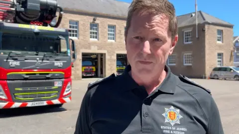 a fire service worker stands in front of a fire truck at the station, speaking to a reporter.