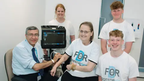 The Robert Jones and Agnes Hunt Orthopaedic Hospital Dr Ayman Askari next to a black scanning machine. He is holding up a scanner to Debs Venezia, who is sitting to his right. Standing behind them is Rachael Griffiths, who has ginger hair. To their right, standing up, is Will Venezia and kneeling below him is Alex Venezia. They are all wearing which tshirts, which read "Do it for Deb"