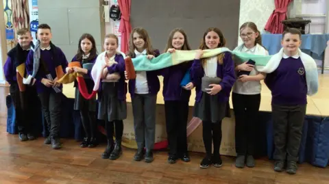 St Giles' CofE Primary School A long knitted scarf held up by nine children wearing blue uniforms in a hall with a wooden floor. The scarf has various colours in it, from red and purple to yellow and green.