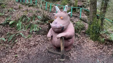 A statue of the Gruffalo , a mythical creature with a large head, orange eyes, a wide month, a round torso, horns, tusks and large claws. The statue is standing in a wooded area with one paw leaning on a wooden post.