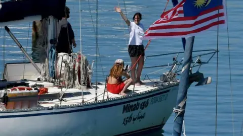 A woman standing on a boat participating in the flotilla waves, as a second woman sits on the deck (April 2026)