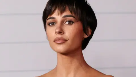 Reuters Naomi Scott olha para a câmera. Ela tem cabelo curto e escuro, olhos castanhos e usa maquiagem neutra.