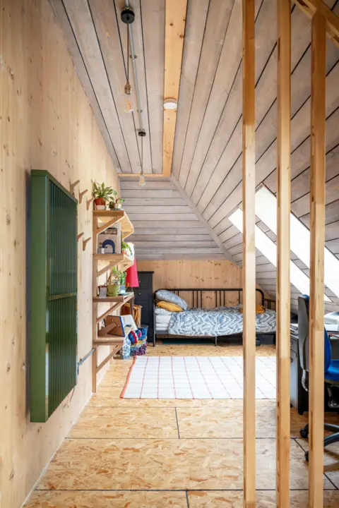 IWC Media More timber in a view through an opening into a bedroom, lined in wood, with a wooden floor and wood shelving. Bright skylights on the sloped right hand side flood the room with light.