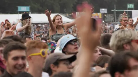 Young Festival goers watching a stage. They are wearing hats and sunglasses. Some girls sit on shoulders. 
