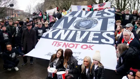 PA Media A group of people hold a large banner which reads 'Lives Not Knives'.