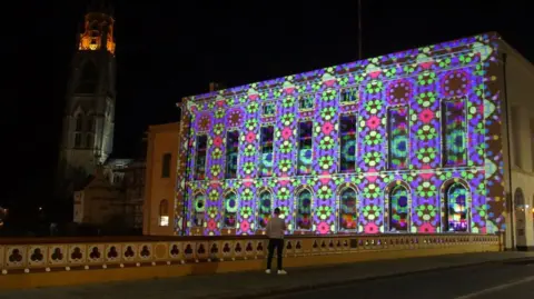 A man standing on a bridge looking at a brightly coloured display projected on to the rear of the Boston Assembly Rooms building, with the tall tower of St Botolph's Church in the background.