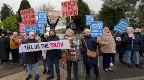 BBC A group of protesters all wearing winter-appropriate clothing. Many are holding protest signs with slogans such as "hands off our Goole hospital". A banner reads "tell us the truth". They are standing on a path in front of a group of tall trees. The sky is overcast.