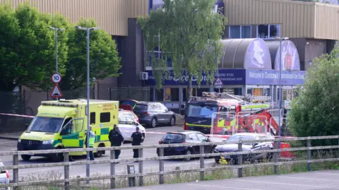 Pacemaker A red and yellow fire engine is parked outside the main entrance of Dundonald Ice Bowl. A green and yellow ambulance is parked close by. A number of cars are parked around the building. A red and white cordon is in place and three police officers stand beside it.