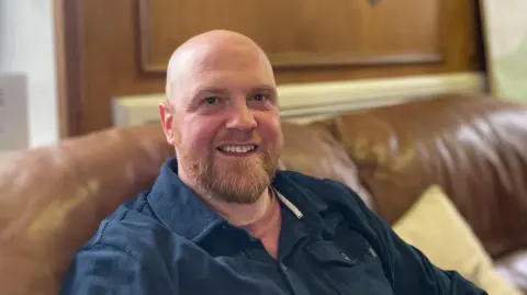 Dave Southall, a man in his 40s, sitting on a brown leather sofa. He is wearing a dark blue shirt and t-shirt. He is bald, has a beard and is smiling.