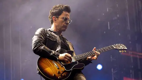 Stereophonics' Kelly Jones on stage, playing guitar. He is wearing a black leather jacket and a pair of glasses.