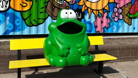 A green bin shaped like a frog placed upon a yellow bench.