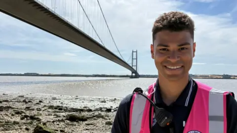 Jake Zuckerman/BBC Yusef Forster standing on Hessle Foreshore with the Humber Bridge visible over his right shoulder. He is wearing a pink hi-vis top and a black polo shirt. He has dark brown hair and is smiling at the camera.