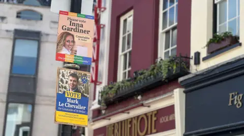 Two campaign posters are attached to a black lamppost on the street. The upper poster reads “Please vote Inna Gardiner, Constable St Helier” with a portrait and a slogan about community, while a second poster below promotes “Mark Le Chevalier for Constable of St Helier.” In the background are multi‑story buildings with red and cream facades, windows with flower boxes, and signs.