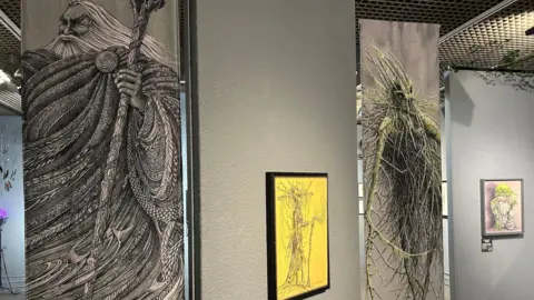 BBC Intricate illustrations of Manx figures, with a large drawing of a man with long flowing white hair a beard and holding a staff.