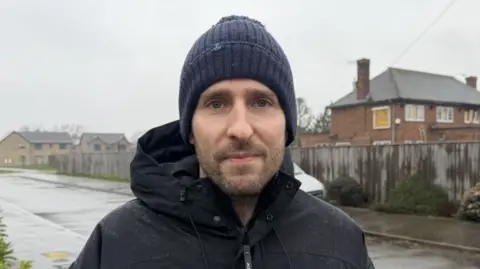 BBC/Hannah Lucas A man looking at the camera. He is wearing a blue bobble hat and a black waterproof jacket. He's standing on a path. There is a fire-damaged building with Georgian-style windows on the right.