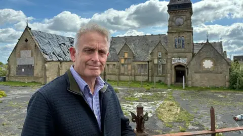 An older man stands in front of a Grade II listed school building made of light coloured stone, in a derelict state. The building is one-storey with a clocktower. Large windows are boarded up, and a extensive playground area is covered in grass and weeds. The man has short white hair, and wears a blue jacket and a blue and white striped shirt.