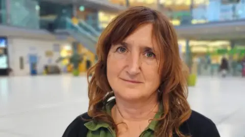 Paul Moseley/BBC Lucy Galvin looks directly at the camera, unsmiling. She has brown hair and is wearing a green top with dark cardigan. She is standing in a glass atrium. 