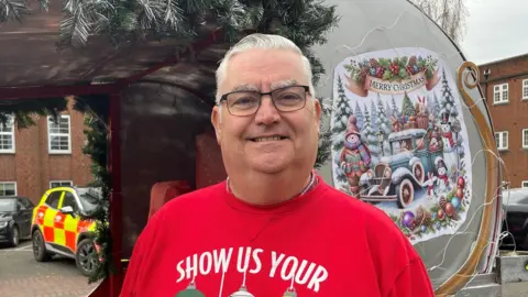 BBC Man wearing a red Christmas jumper stood in front of a sleigh