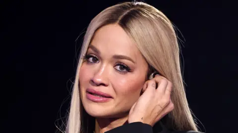 Rita Ora holds her earpiece as she looks emotional paying tribute to Liam Payne