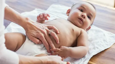 Getty Images A baby on its back on a muslin having its disposable nappy changed. A woman's hand can be seen resting on its belly while her other hand is opening the nappy fastening on the side. 