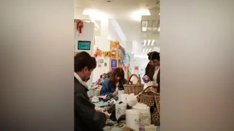 Love Saves the Day Three people - two women and a man - at a table covered in oddments such as pieces of fabric, sewing baskets and tubs of sewing materials. They are in a large hall with overhead stripped lighting and bunting of multicoloured material