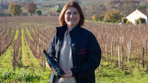 Coralie de Bouard staat in een wijngaard met een fles wijn in haar hand