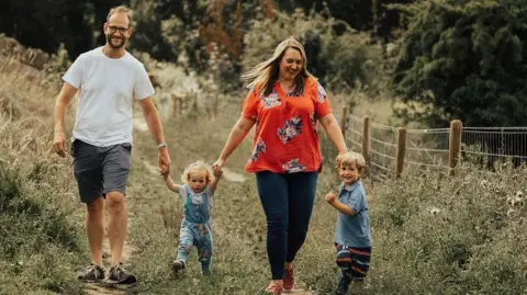Family handout A man and woman holding the hands of their two children, a young girl and boy, walking along a grass path. The man is wearing a white top and grey shorts with trainers and glasses and smiling at the camera. A little girl is wearing blue dungarees. A woman is looking down at her little boy and has long blonde hair and an orange flowery top and blue jeans. The little boy is standing to the side and smiling at the camera.