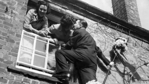 Mirrorpix via Getty Images A black and white image of a Tutti man climbing a ladder to get the traditional kiss the window during the Hocktide festival at Hungerford in April 1955. Two women are leaning out of the house's window. One of them is kissing the Tutti man. The other one is smiling at the camera.