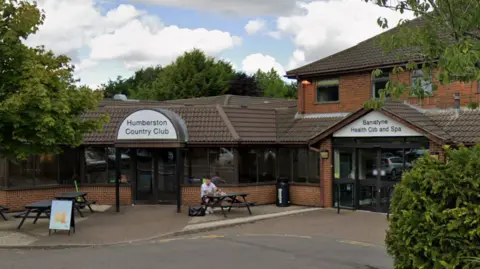 The entrance to Humberston Country Club. A man is sitting on a black bench outside of the entrance. They sky is blue with white clouds. 