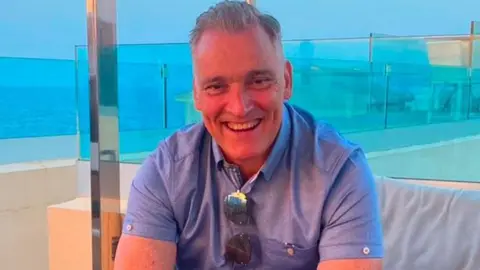 Stephen Howes smiling towards the camera. He is wearing a blue T-shirt, with sunglasses hanging down having been hooked in the his shirt which is open at the collar. 