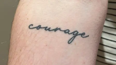 Rachel Carlson A woman is holding up her right arm.  She has a tattoo reading "courage" just below her elbow joint