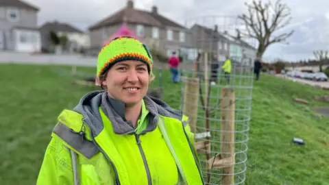A woman in a high-viz jacket stands in front of newly planted tree on an area of green space in Whitely, Plymouth. There are other people planting more trees in the background. You can see semi detached houses in the background. 
