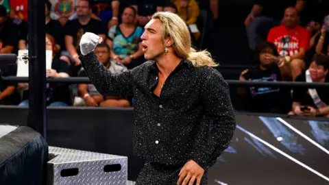 Getty A wrestler, with long blonde hair in a ponytail, wearing a black glittery suit and one white glove fist pumps