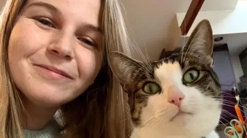 Laura Taylor A woman takes a selfie style image of herself and her cat that sits next to her. She has long blonde hair and is smiling. The cat has some tabby markings around its eyes as well as white fur around its nose and mouth. 