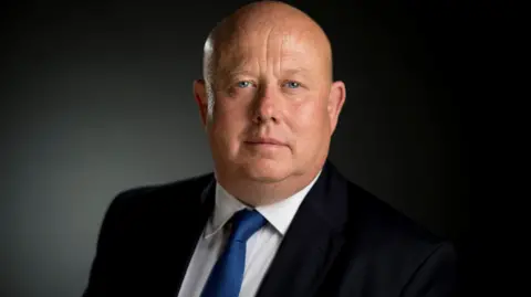 Former council leader Ian Hudspeth. He is a bald man with blue eyes wearing a black suit, white shirt and blue tie - he is looking at the camera.
