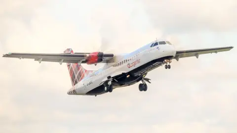 A propeller passenger plane in flight with its wheels down. The aircrfat has Loganair livery, including a red tartan tail.