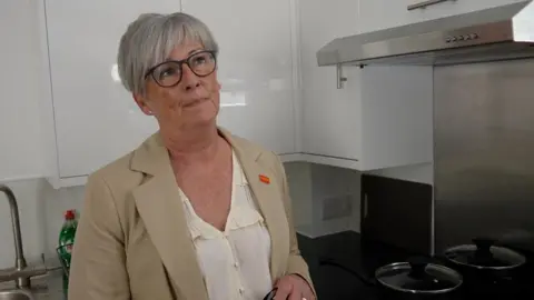A woman wearing a blazer is in a newly refurbished flat for young people , behind her is a brand new kitchen with new cooking pans.