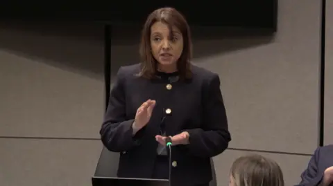 Senedd Cymru Shavanah Taj, a woman with shoulder length brown hair and a dark jacket, stands up in a room and is speaking.