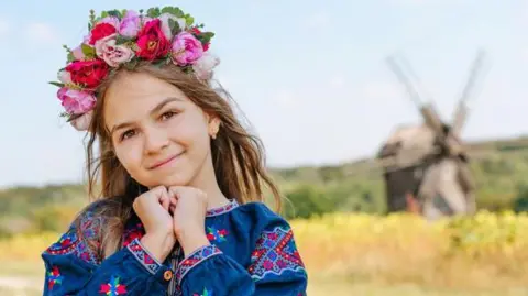 A girl wears Ukrainian national dress and a Vinok wreath of flowers outdoors. Her dress is blue with brightly-coloured patterns and the wreath is pink, red and purple.